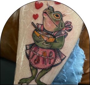 A fresh tattoo of a cute frog doing some baking, that is covered in a second skin bandage.