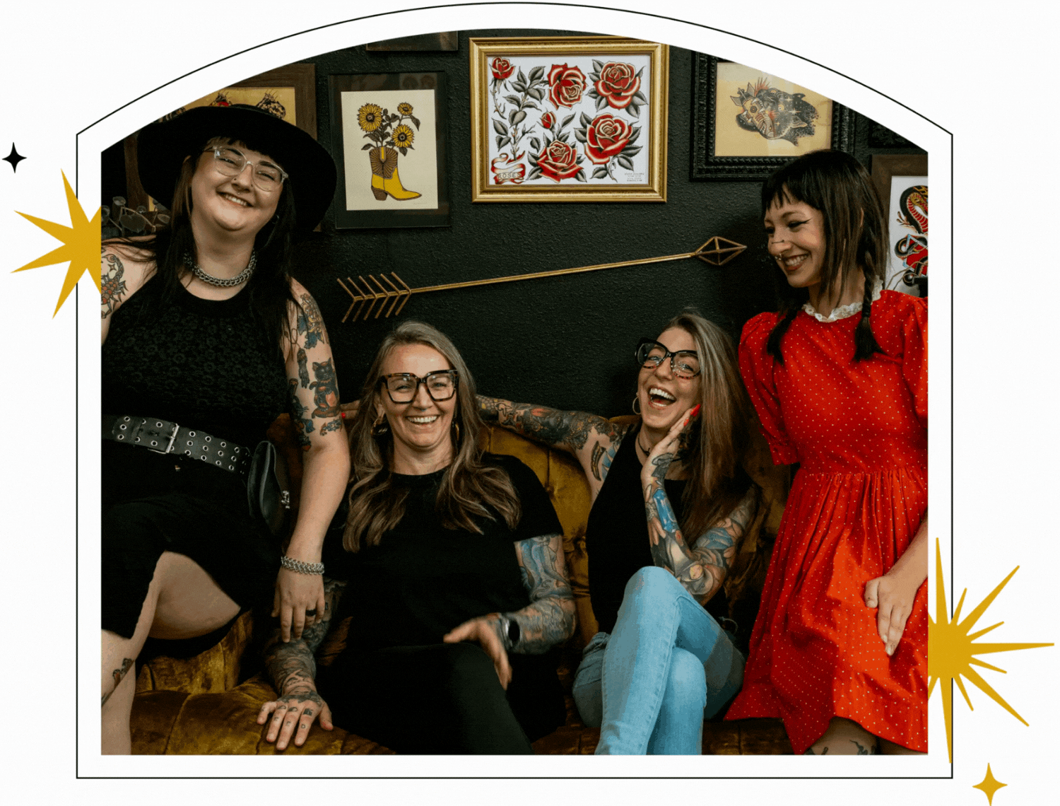 Four smiling women with tattoos, the resident artists of Black Magic Tattoo, sit together on a gold couch in front of framed artwork.