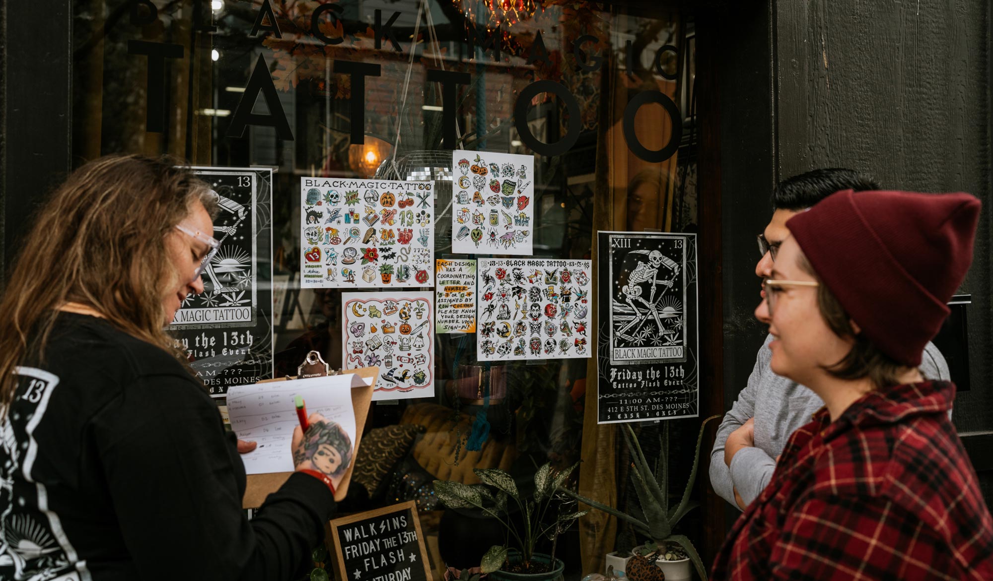 Clients outside of Black Magic Tattoo picking tattoo designs taped to the window while Ashley writes them down on a clipboard.